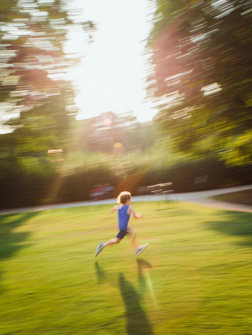 A still from a Super 8 home movie, showing a child running through a park on a brightly lit summer afternoon. The image exhibits the characteristic traits of Super 8 film, including noticeable film grain, flickering light, and slightly faded colors. The footage is slightly overexposed with a warm color temperature, imbuing the scene with a nostalgic and dreamlike quality.