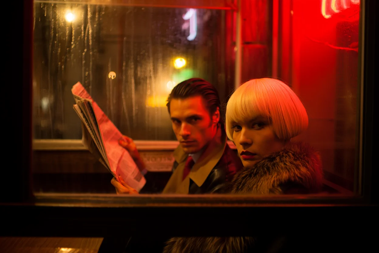 A cinematic night scene from inside a diner, framed by a rain-streaked window, featuring two individuals peering in from the street. The shot is a medium close-up from a low angle, creating a voyeuristic and suspenseful mood. The left subject, with slicked-back hair and a trench coat, holds a newspaper, partially obscuring their face. The right subject, a woman with a platinum blonde bob and a fur collar, peers over his shoulder. The background is a blur of warm diner light and neon signs reflected on the wet pavement, with a dominant red glow. The shallow depth of field focuses on the intrigue of the subjects' expressions against the cinematic backdrop. High quality film noir style