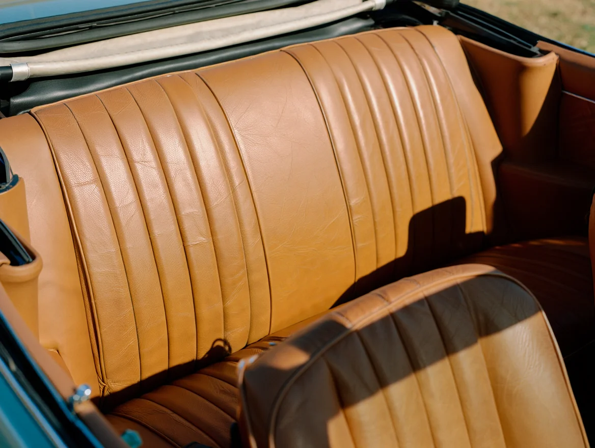 A vintage film photograph taken from inside a convertible car shows the tan leather backseat with visible stitching and weathered creases. The folded fabric roof is partially visible above, creating a natural frame around the composition. Warm sunlight illuminates the worn leather surfaces, highlighting the authentic patina and texture of the material. The backseat spans the full width of the frame, with subtle shadows cast by the interior components. Vintage 35mm film photography with natural grain texture and soft ambient lighting.