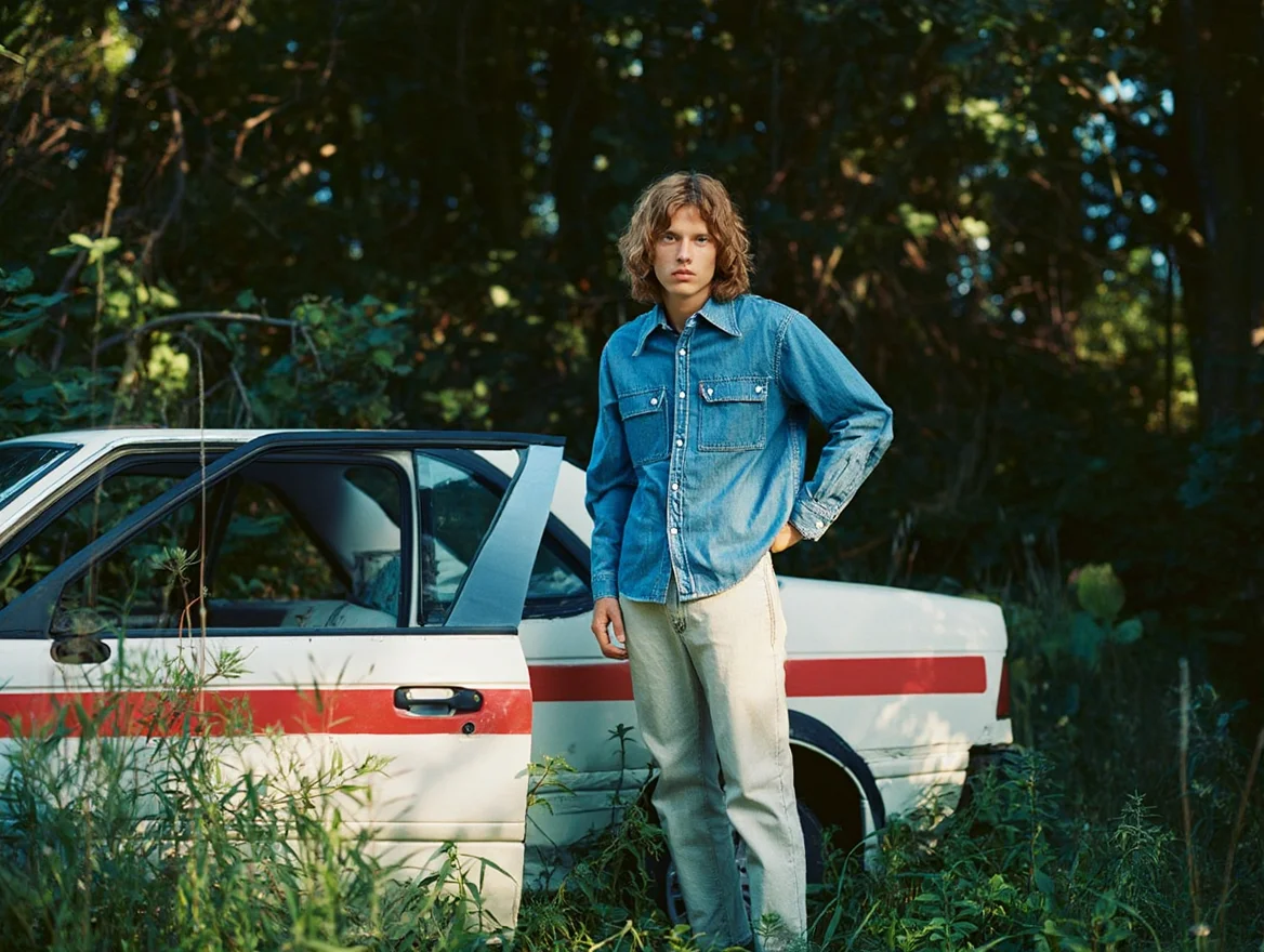 A casual snapshot shows a figure wearing a denim shirt and light-colored pants standing with one hand on hip next to a partially visible white car with red striping, the car appearing wrecked or abandoned among overgrown brush. Natural daylight filters through trees in the background, casting dappled shadows across the scene. Wild grass and foliage grow in the foreground around the car. The photograph exhibits characteristic 1990s film qualities including muted colors with blue-green color shifts in shadow areas, subtle film grain texture, and slightly soft focus throughout the frame. Amateur film photography from the 1990s with natural lighting and characteristic film color rendering.