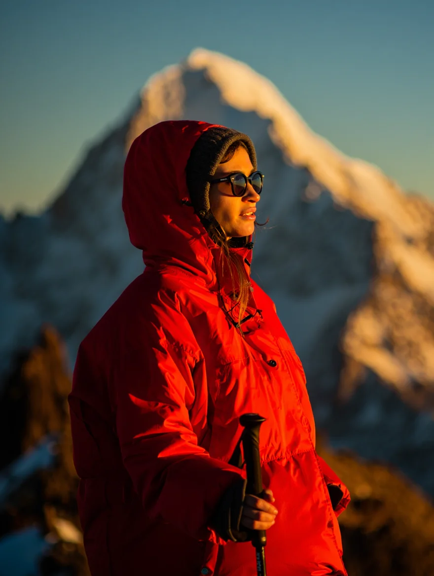 A lone female hiker pauses on a ridge line in the Andes, photographed in a medium shot with a snow-capped mountain peak creating a dramatic backdrop. Bathed in the golden light of the setting sun, she stands facing the camera, wearing a bright red parka and sunglasses, while holding a trekking pole. The image exhibits a warm color palette with a cinematic feel.